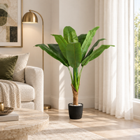 100 cm artificial banana tree with broad green leaves and a single trunk in a black pot beside a sofa and sunlit window.