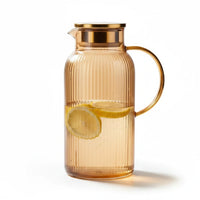 Trendiest amber ribbed glass pitcher with gold lid and handle containing water and lemon slices on white background