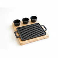 Trendiest cast-iron grill plate with wooden serving board and sauce bowls, close-up product view.