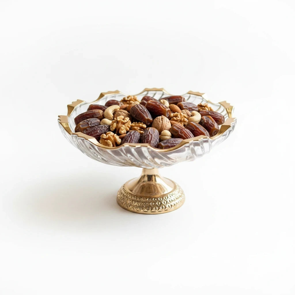 Trendiest decorative glass pedestal bowl with gold rim filled with assorted nuts and dates on a white background