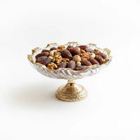 Trendiest decorative glass pedestal bowl with gold rim filled with assorted nuts and dates on a white background