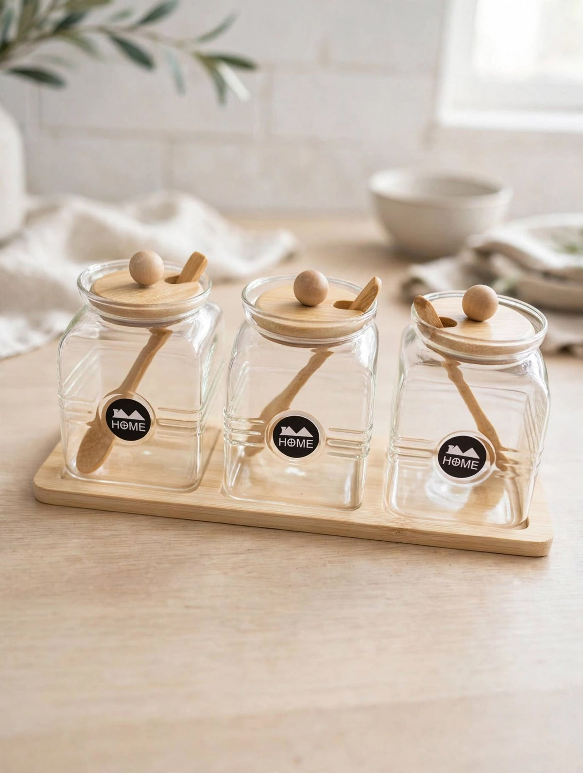 Trendiest glass spice jar set of three with bamboo lids, wooden spoons, and a display tray on a kitchen counter.