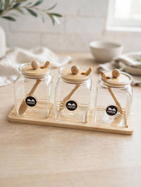 Trendiest glass spice jar set of three with bamboo lids, wooden spoons, and a display tray on a kitchen counter.