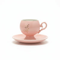 Trendiest pink ceramic teacup with a pedestal base, gold floral detail, and matching saucer on a white background.
