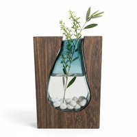 Trendiest handcrafted dark wood vase with a blue-tinted hand-blown glass insert containing white pebbles and greenery.